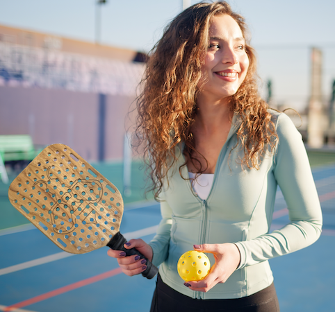 Woman holding a Bee Quiet noise reducing harwood pickleball paddle and a ball on a court
